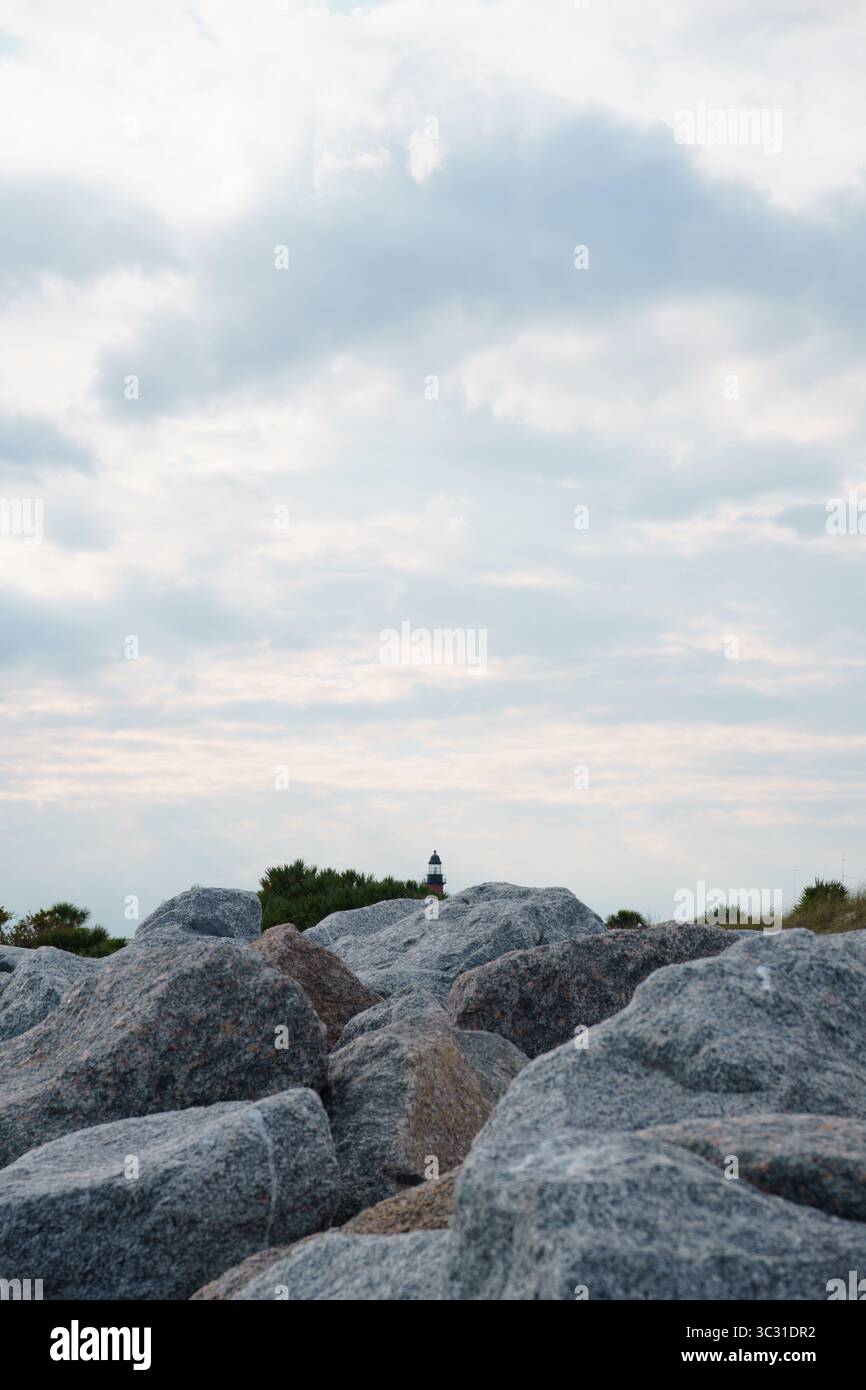 Faro distante visibile sopra grandi rocce costiere sotto un cielo nuvoloso. Foto Stock
