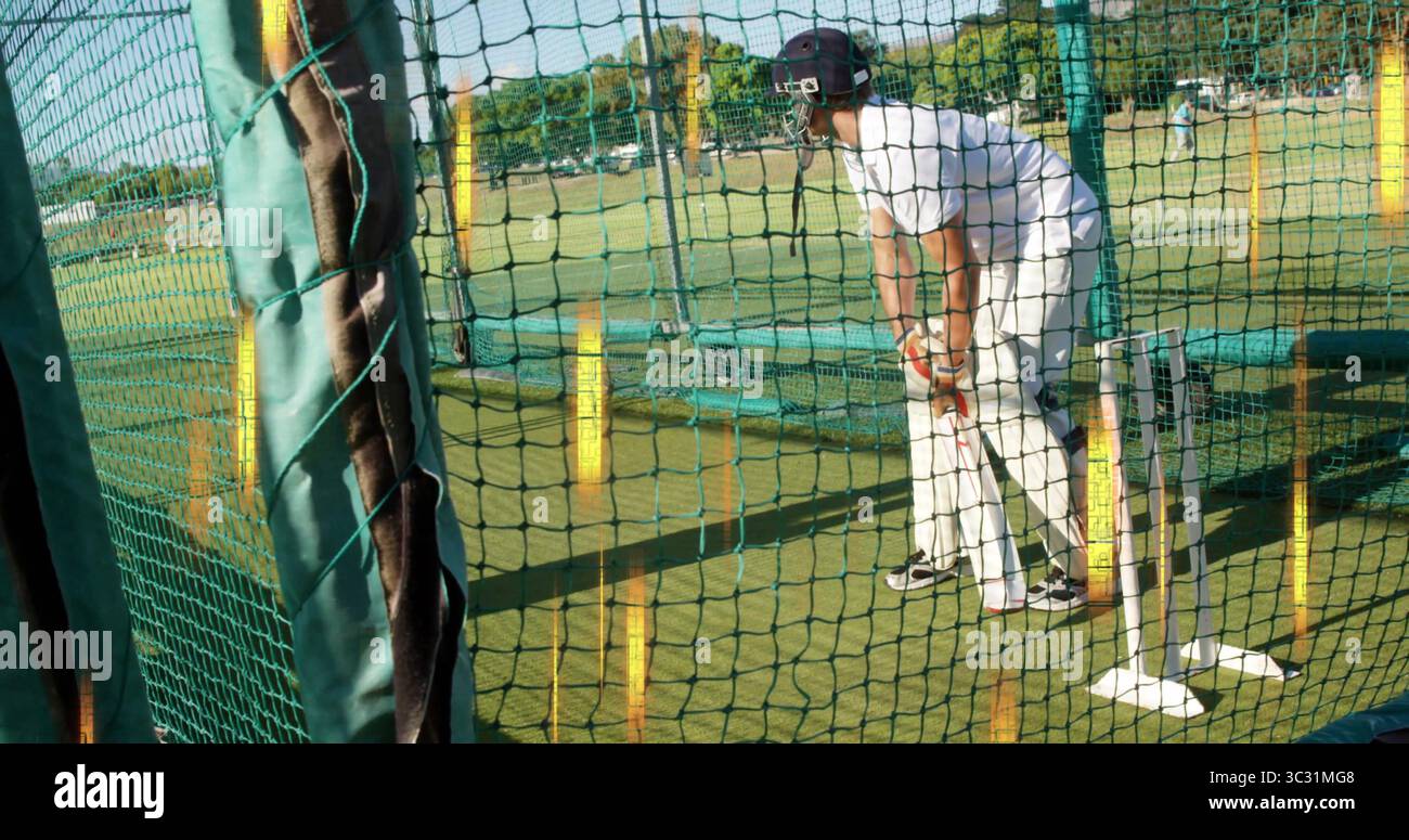 Praticare il cricket maschio battitore in uniforme bianca tenendo la mazza alla gabbia di rete sul campo, con ceppi Foto Stock