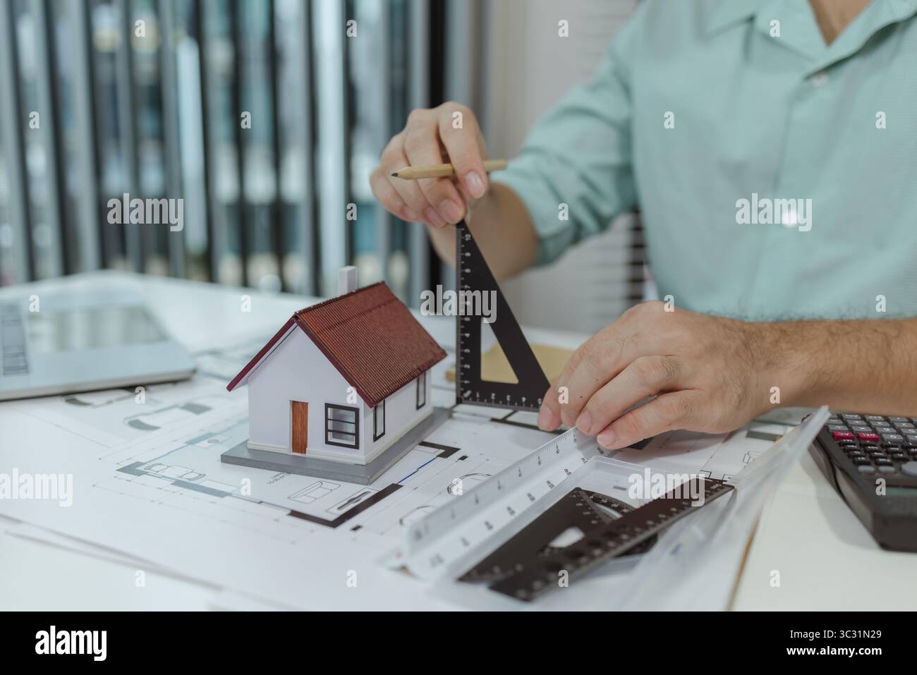 Un architetto utilizza un righello e una matita triangolari per misurare un modello di casa posizionato su progetti architettonici, controllando proporzioni e dettagli durante t Foto Stock