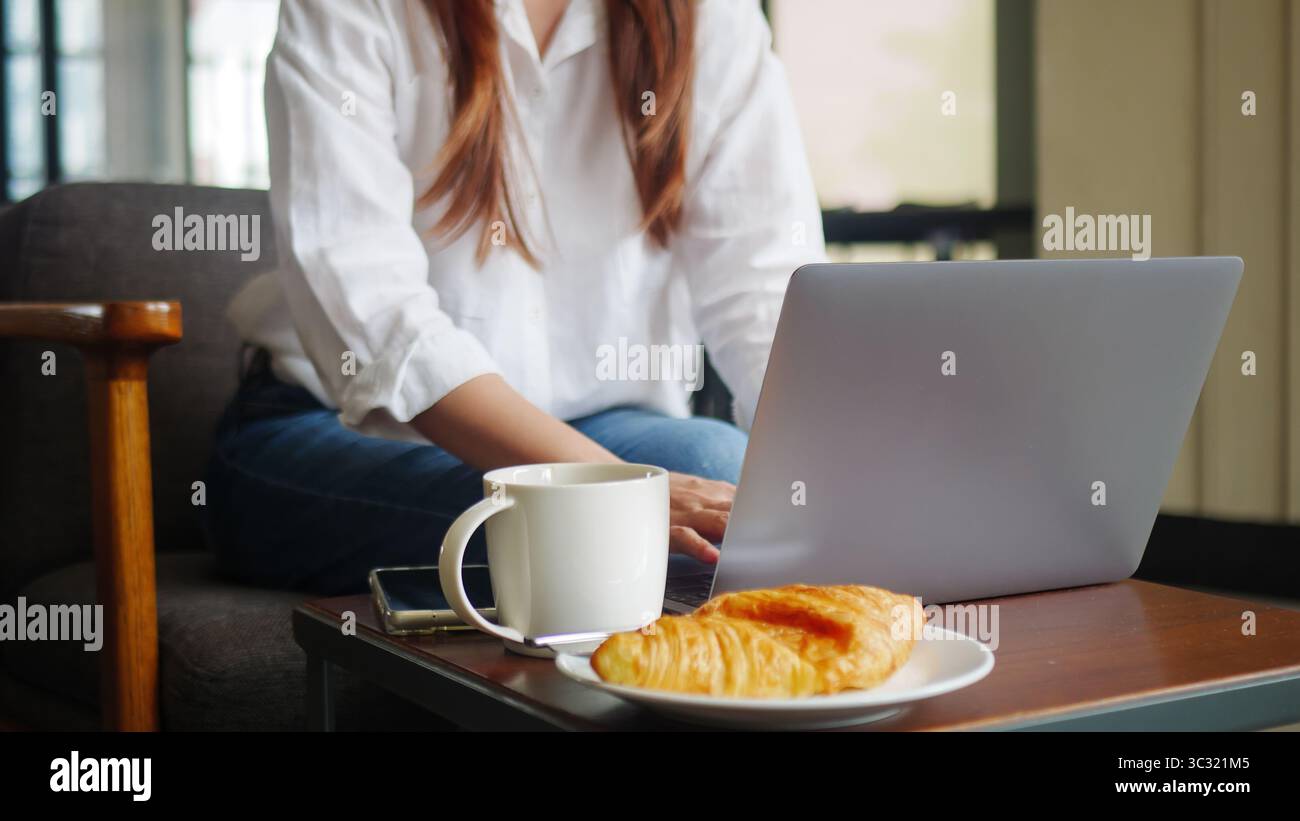 vista frontale della donna che utilizza un computer portatile, lavora da casa al mattino con caffè caldo, panetteria con croissant e telefono cellulare sul tavolo. anonimo Foto Stock