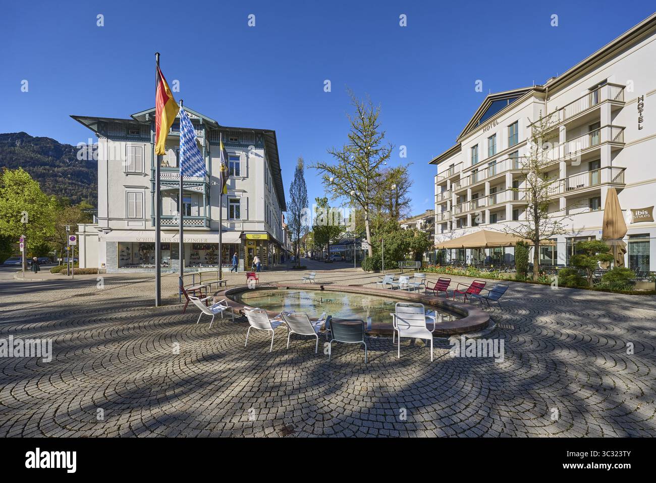 Wisbacherbrunnen, fontana, sedie pubbliche, paletti, bandiere, bandiera tedesca, bandiera bavarese, edificio, hotel, ibis Styles, alberi, cielo blu senza nuvole, Ludw Foto Stock
