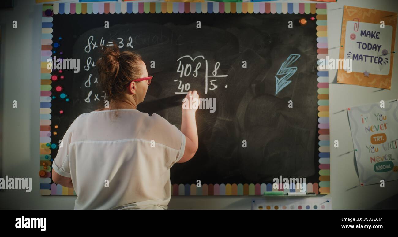 Vista posteriore dell'insegnante femminile che scrive sulla lavagna, spiegando la moltiplicazione e la divisione ai bambini delle scuole elementari durante la lezione di matematica. Corso di matematica in classe moderna. Colpo statico. Foto Stock