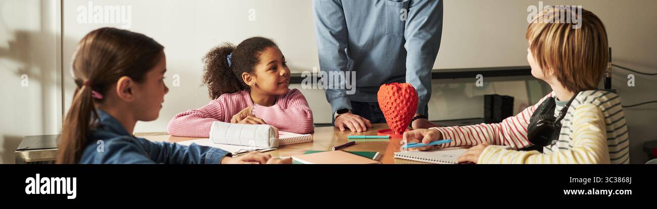 Intestazione di un gruppo di bambini diversi seduti a tavola ascoltando un insegnante maschio caucasico che spiega la lezione, materiale scolastico sparsi sulla scrivania, studenti che mostrano espressioni attente Foto Stock
