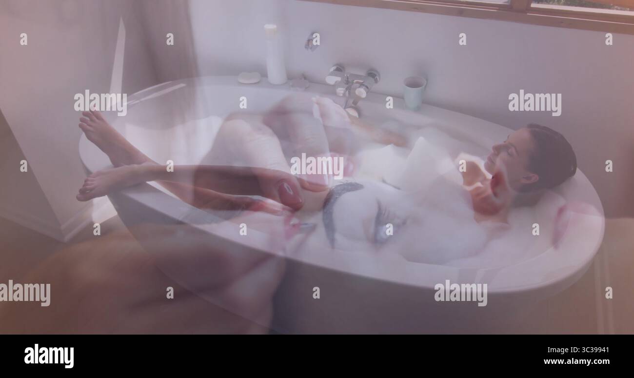 Donna sdraiata con i capelli tirati indietro e immersa nella vasca da bagno del bagno di casa, schiuma, rivestimento dello smartphone Foto Stock