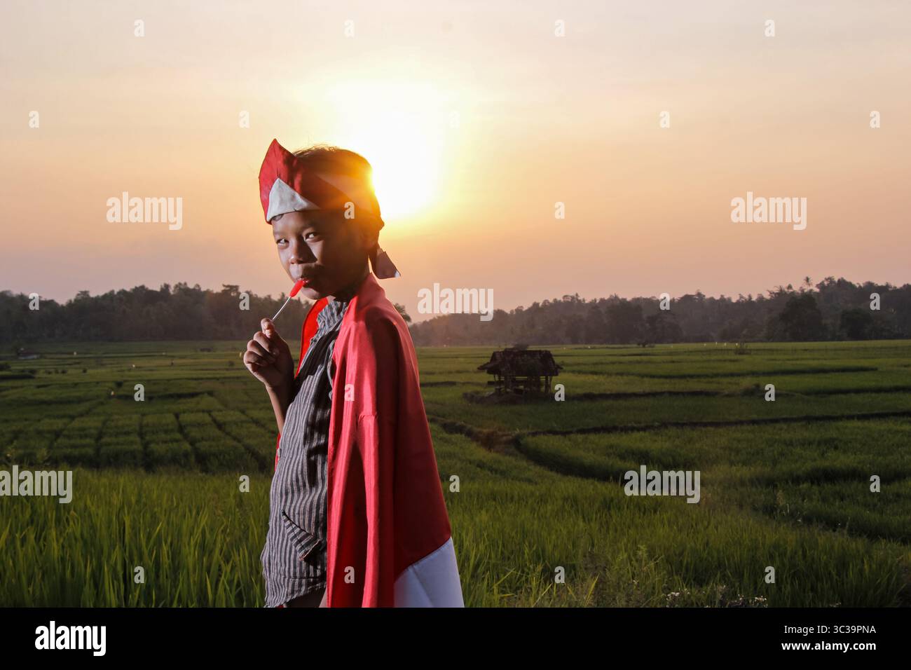 Un ragazzo indonesiano sorride mentre mangia caramelle nel mezzo di un campo di riso con un bellissimo tramonto, lo spirito della giornata indonesiana Foto Stock