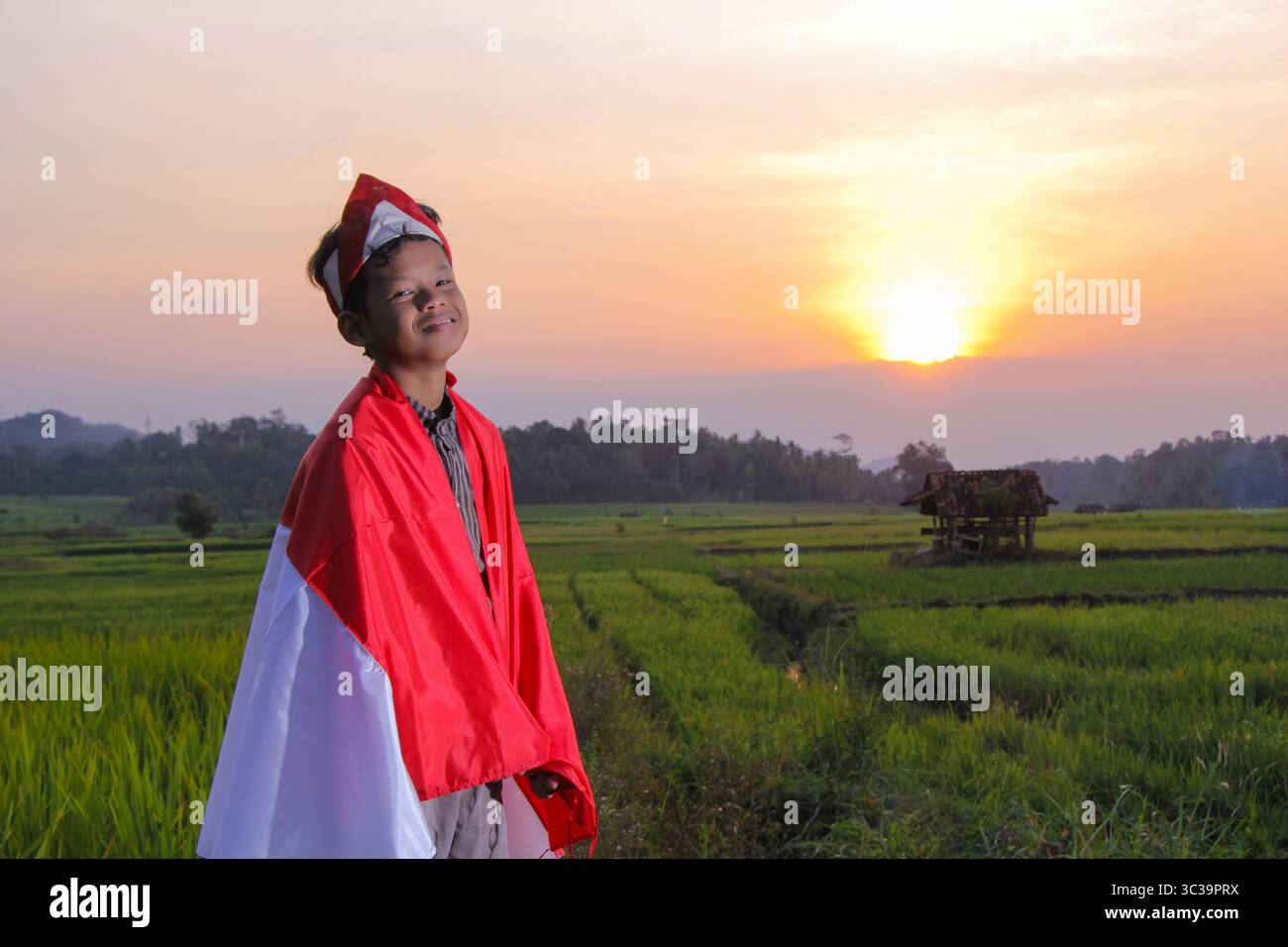 Indonesiano Boy è orgoglioso di alzare Flag mentre il sole tramonta su campi di riso e capanne sullo sfondo Foto Stock