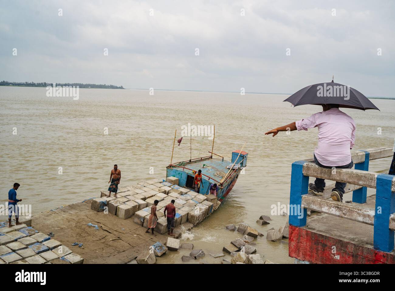 Un uomo dirige i lavoratori che scaricano blocchi di cemento da una barca su una riva del fiume in Bangladesh, mostrando lavoro in riva al fiume, lavoro di squadra e vita. Foto Stock