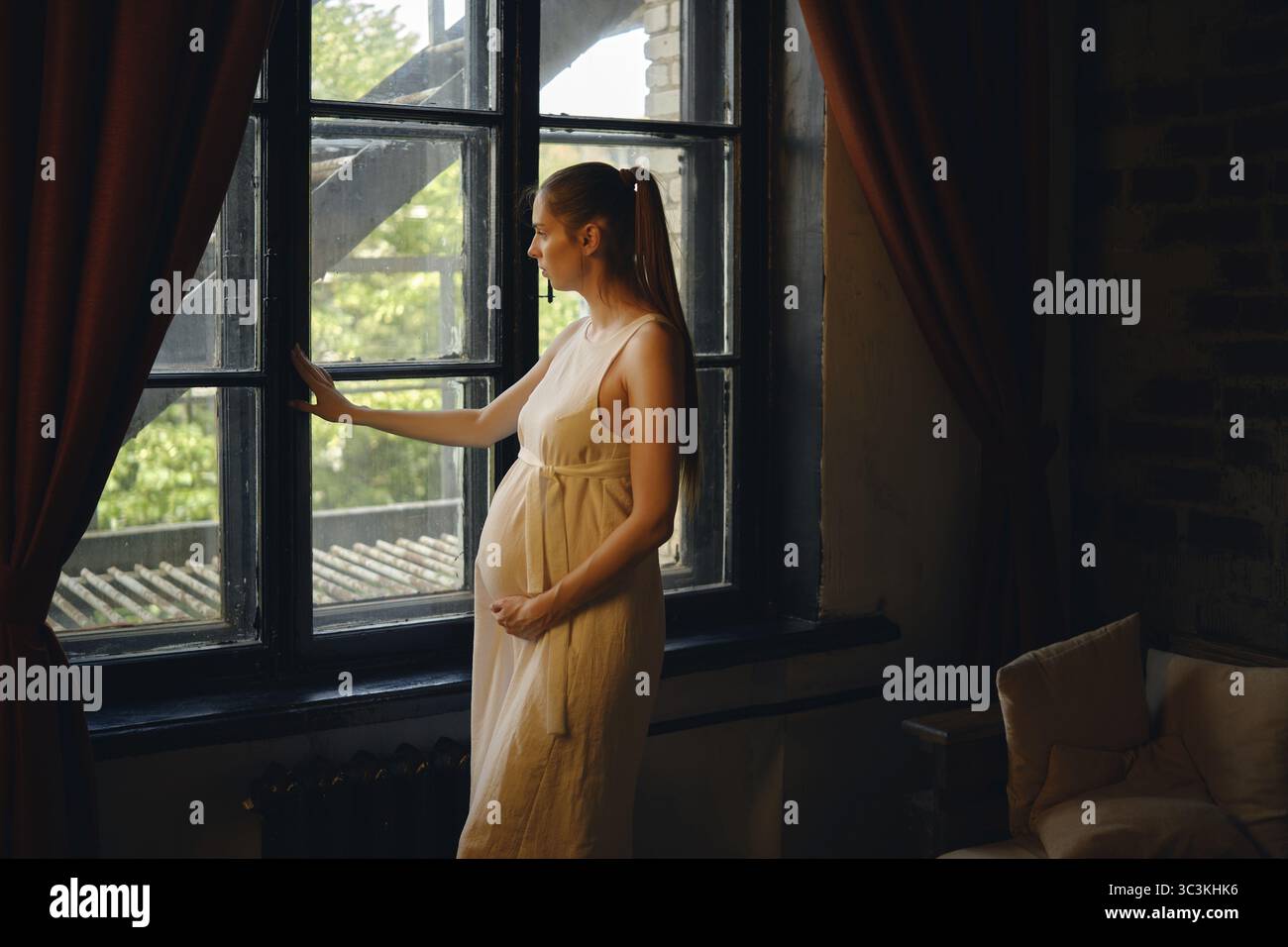 Una donna incinta sta accanto a una finestra nella sua casa, ammirando la vista dall'esterno. Appare riflessiva e serena, vestita con un abito semplice e fluente Foto Stock