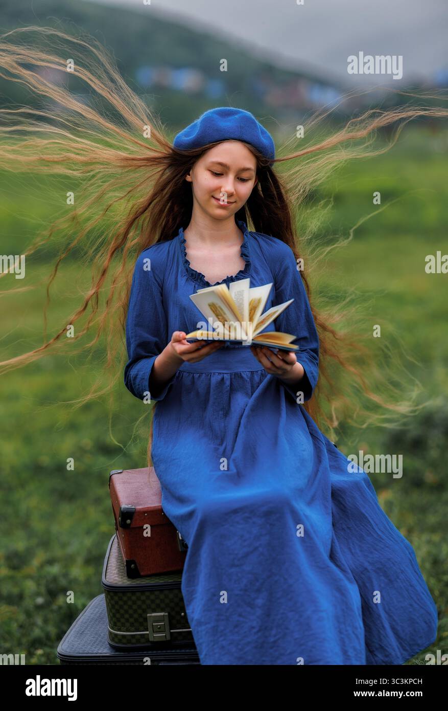 Sognante ragazza d'epoca in abito blu che legge un libro su valigie impilate in campagna sotto il cielo nuvoloso Foto Stock