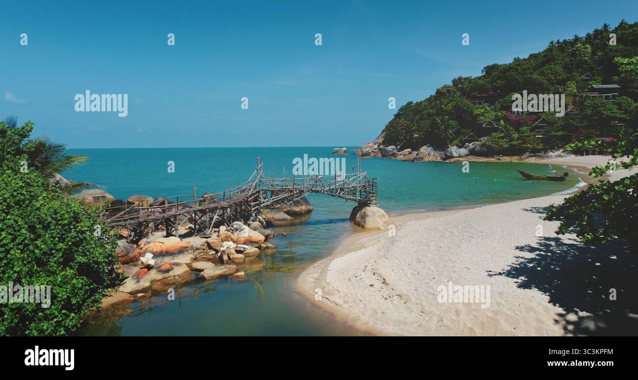 Drone aereo di un ponte di legno ricchissimo che collega rocce su acque turchesi incontaminate, che conduce ad una tranquilla spiaggia di sabbia bianca annidata in una lussureggiante insenatura tropicale in Thailandia Foto Stock