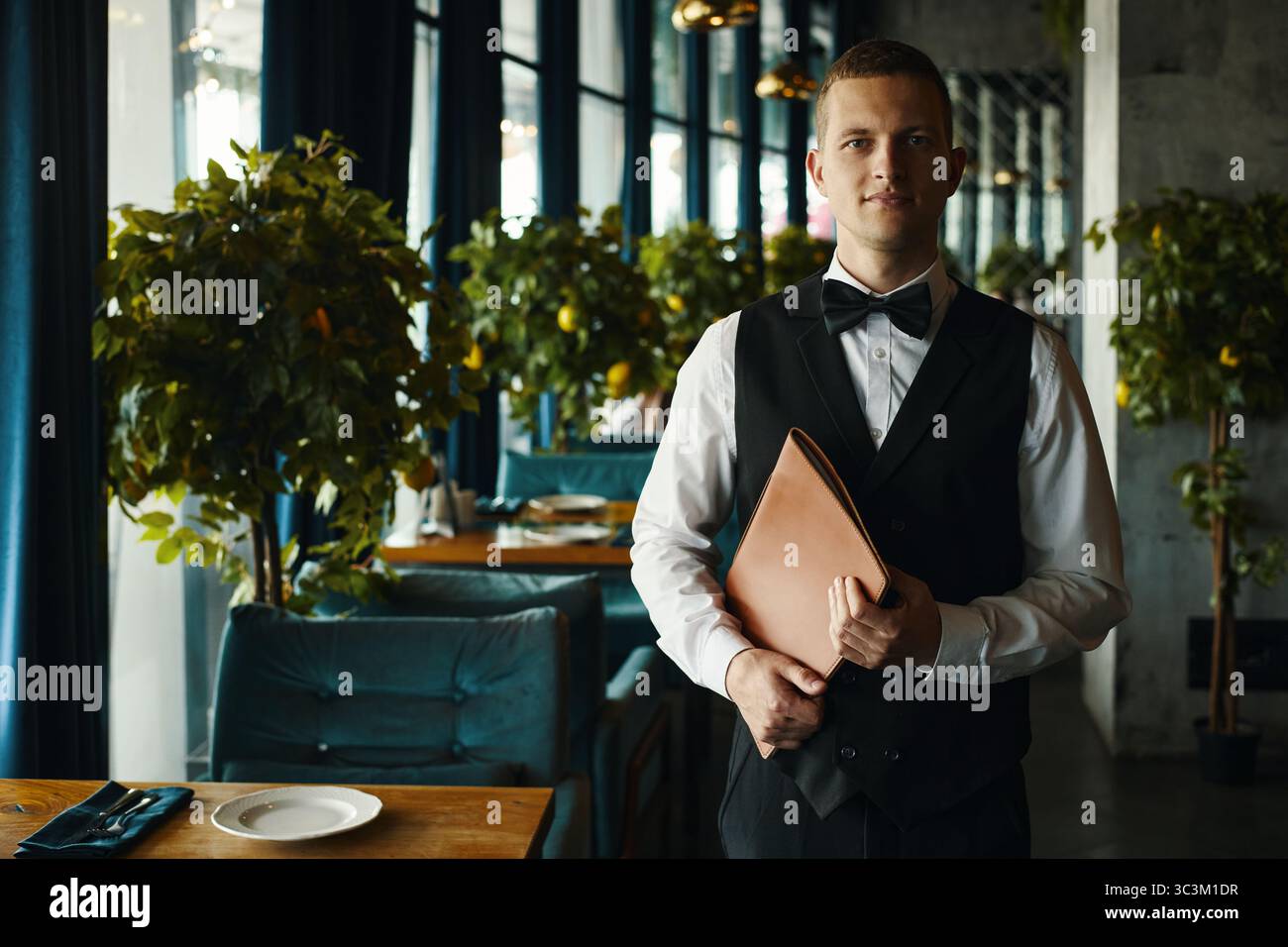 Ritratto di un giovane uomo caucasico adulto in piedi al ristorante, con cartellina del menu che indossa gilet formale e cravatta che guarda la fotocamera con espressione neutra al chiuso Foto Stock