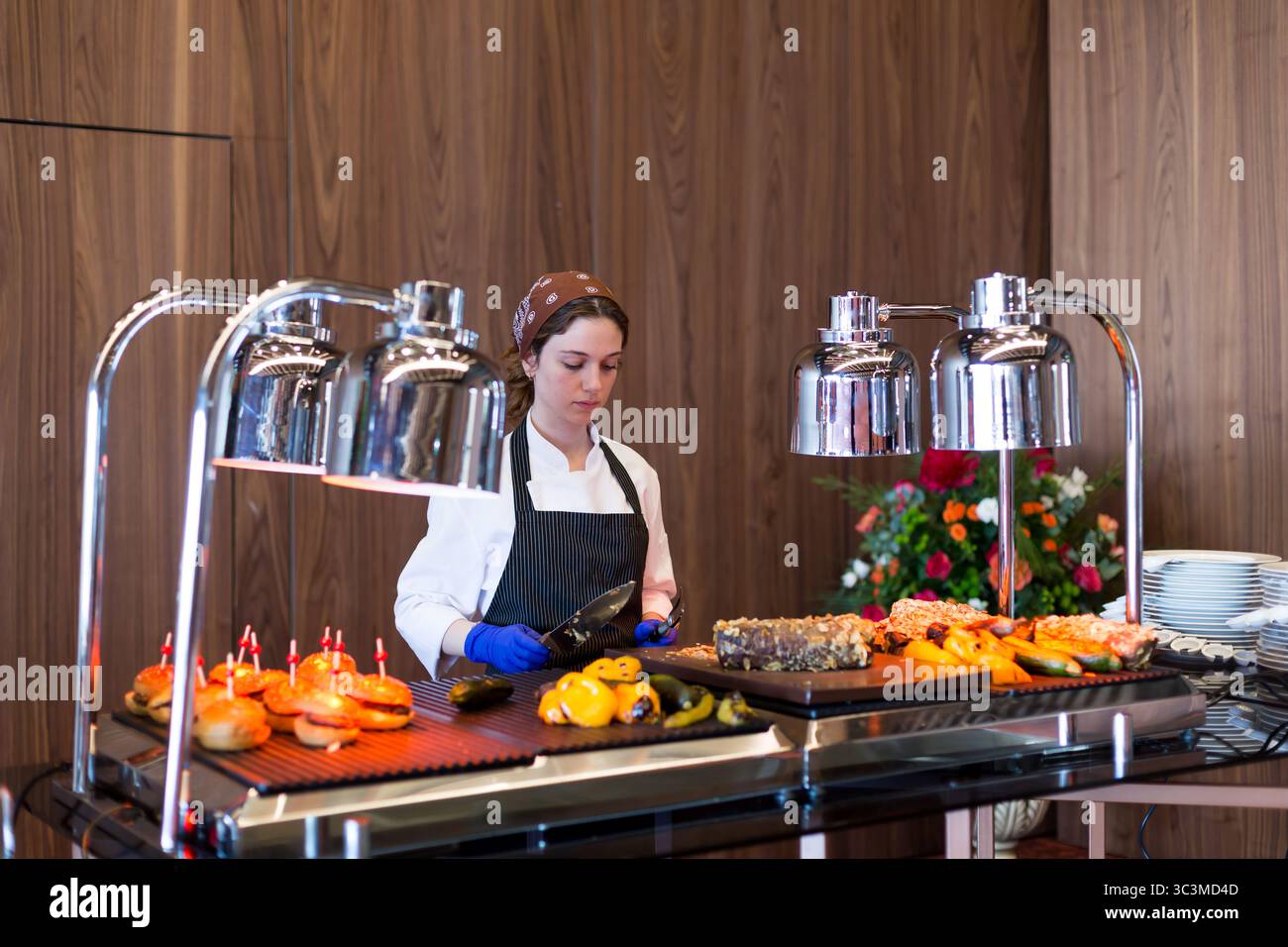 Istanbul, Turchia - 24 giugno 2022: Donna in grembiule e fascia che serve piatti gourmet presso la moderna stazione a buffet con lampade termiche, serve piatti freschi in alto Foto Stock