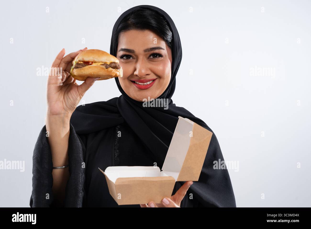 Allegra giovane donna musulmana in un tradizionale abaya nero, con un hamburger e una scatola da asporto, incarna la moderna fusione culturale e la cultura del fast food Foto Stock