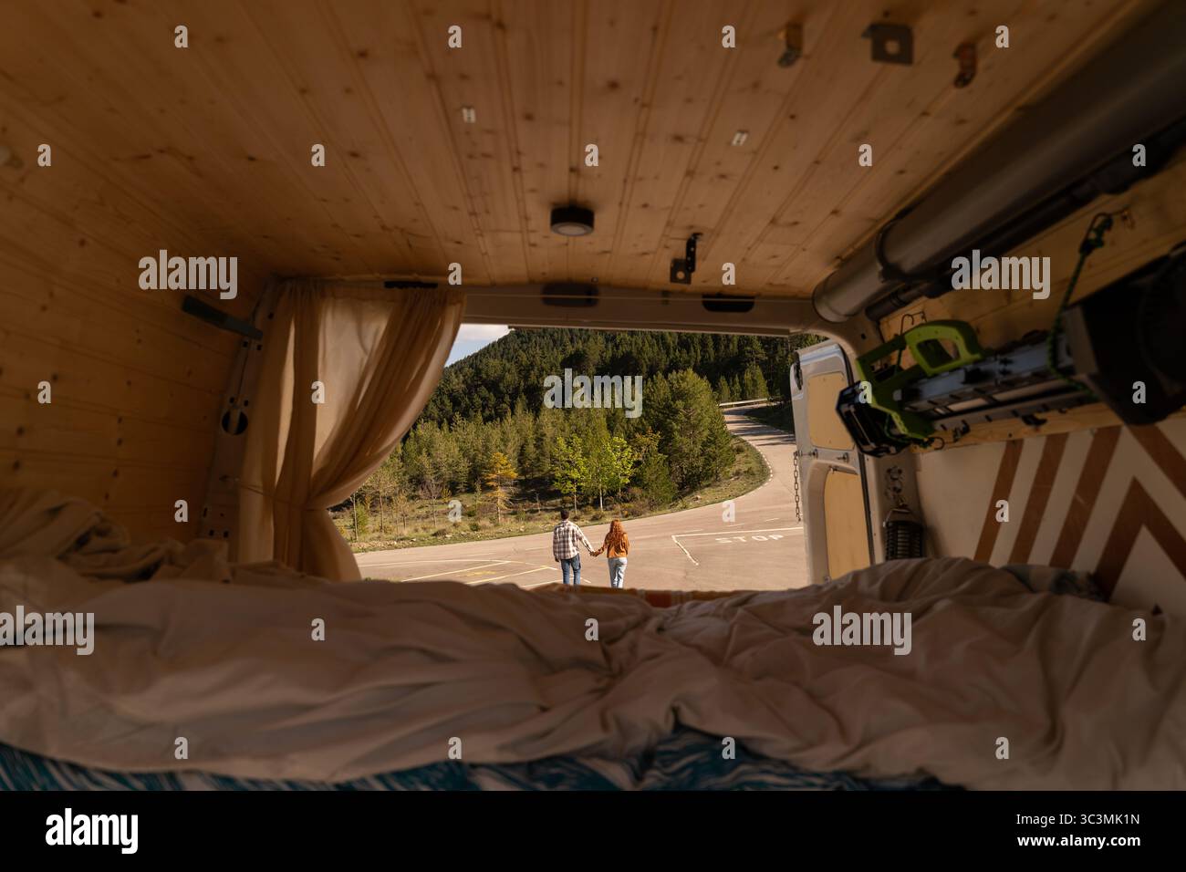 Un confortevole camper interno, visto dal retro, mostra una coppia che tiene per mano un'avventura all'aperto. La lussureggiante foresta verde e la strada tortuosa inv Foto Stock