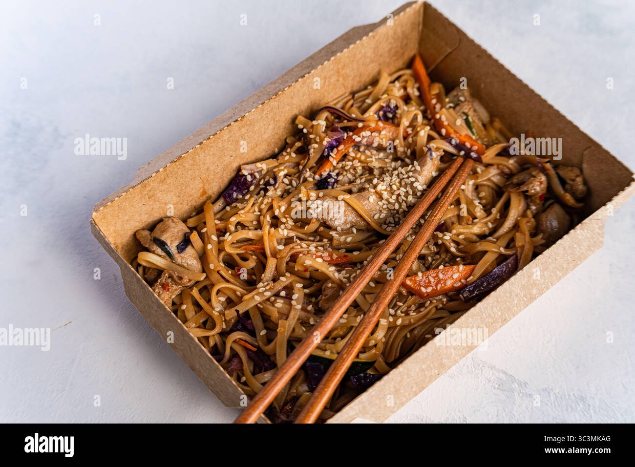 Una deliziosa porzione di spaghetti asiatici fatti a mano conditi con semi di sesamo, carote, cavolo rosso e pollo. Presentati in una scatola di cartone marrone, emphas Foto Stock