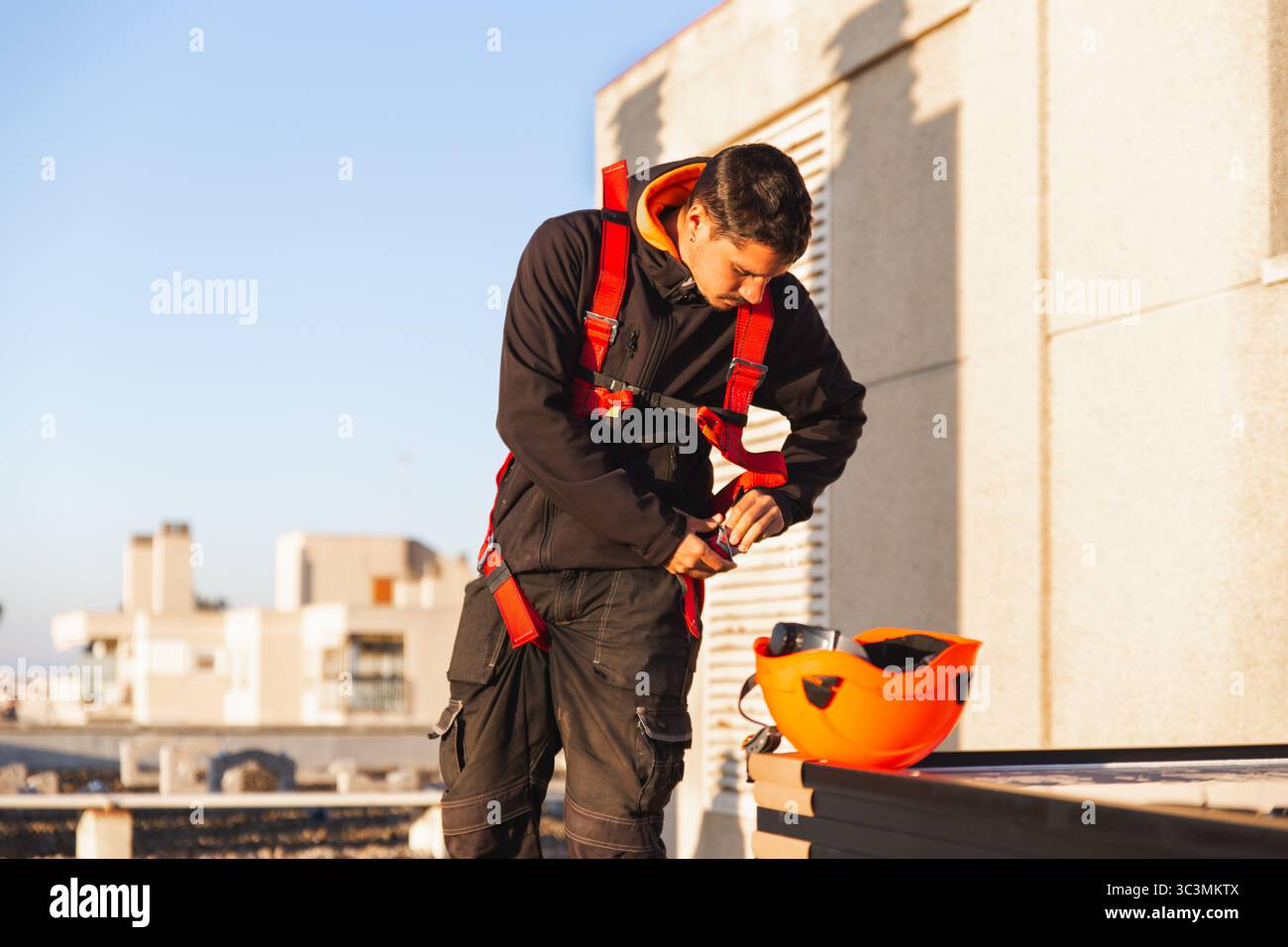 Un operaio esperto in attrezzatura di sicurezza regola un'imbracatura sul tetto, preparandosi a installare pannelli solari. Lo skyline urbano è visibile, evidenziando l'inte Foto Stock