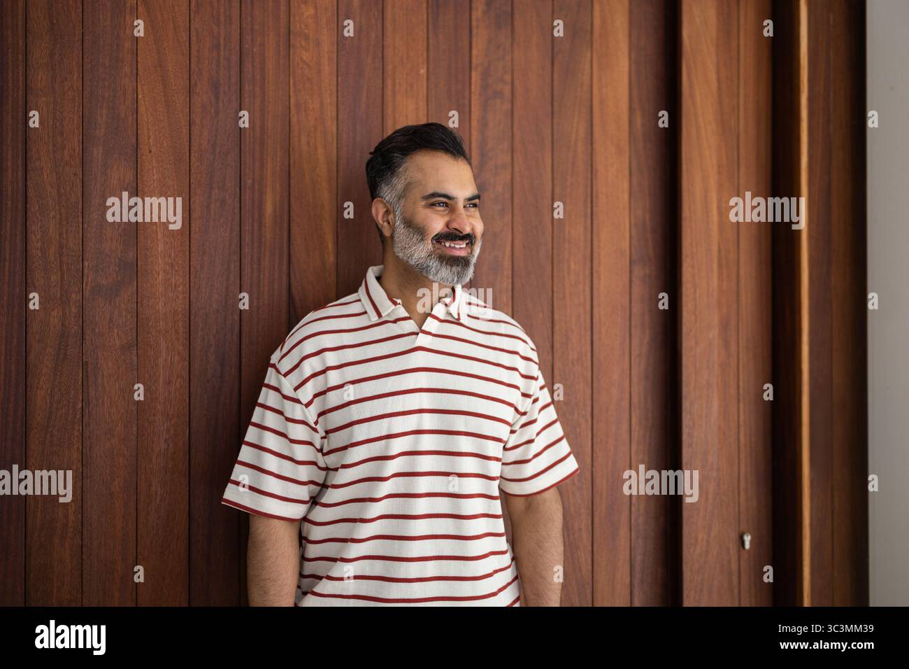 Un uomo indiano sorridente con una camicia a righe si erge contro un muro di legno, godendosi una calda giornata estiva. La scena informale trasmette relax e svago, ideale Foto Stock