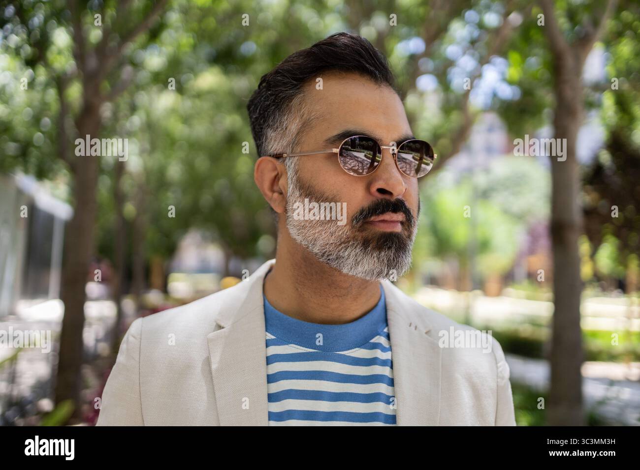 Un uomo indiano che indossa occhiali da sole e un abbigliamento estivo casual si erge tranquillamente all'aperto, godendosi una giornata di sole. Lo sfondo presenta una vegetazione lussureggiante, Foto Stock