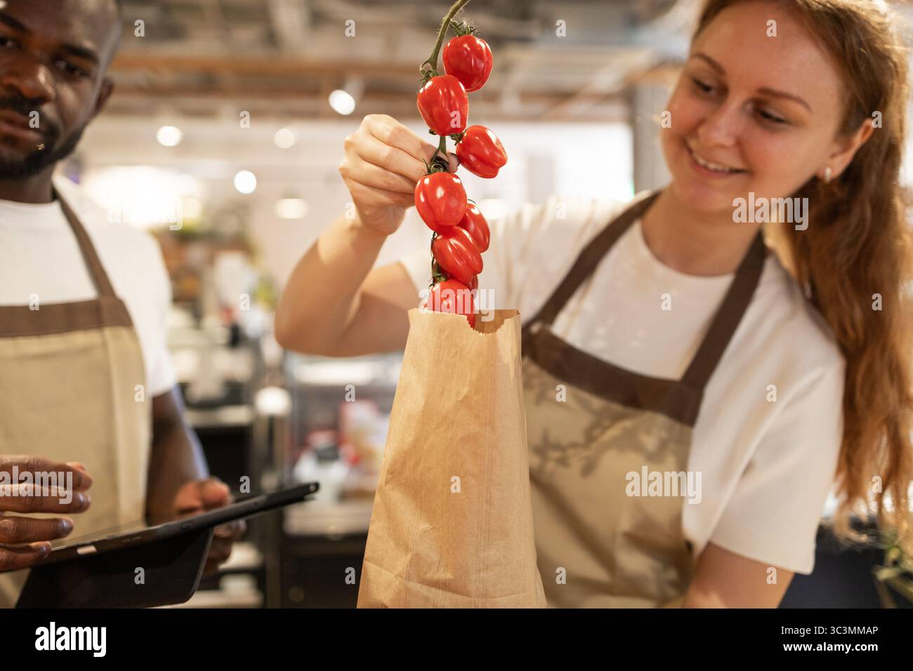 Un uomo e una donna lavorano insieme in un negozio ecologico, confezionando pomodori freschi in un sacchetto di carta. L'atmosfera mette in risalto la sostenibilità e il lavoro di squadra i. Foto Stock
