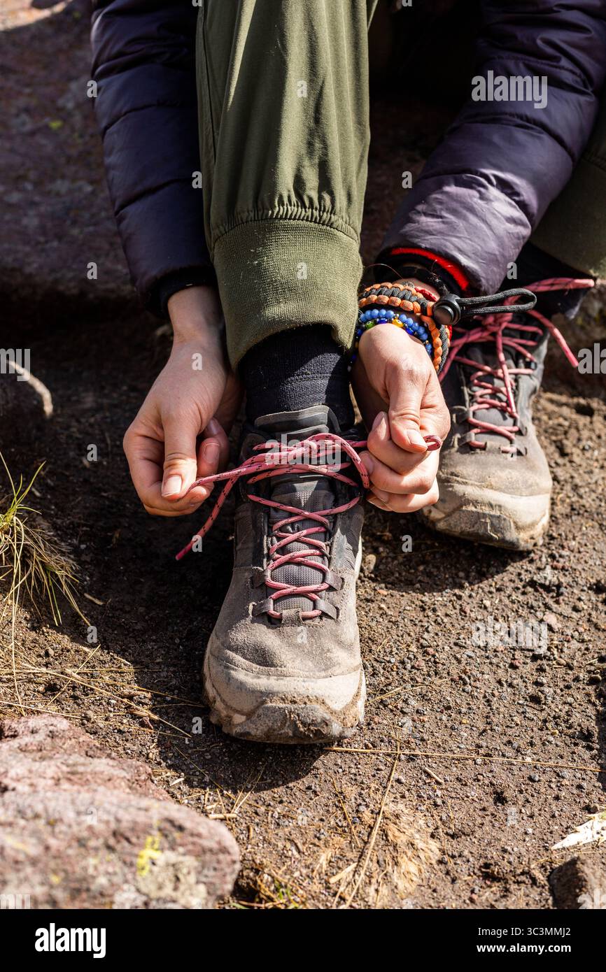 Un escursionista si ferma su un aspro sentiero di montagna per inginocchiarsi e allacciare con cura robusti stivali da trekking, preparandosi per il viaggio che ci attende. La luce solare filtra attraverso Foto Stock