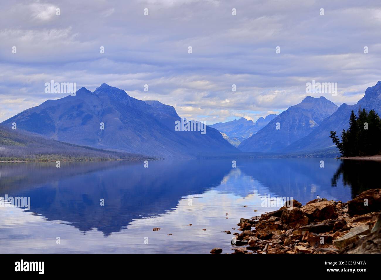 Le maestose cime montuose si innalzano sopra le acque cristalline del lago McDonald, riflettendo le spettacolari nuvole e il litorale sotto un cielo lunare nel Glacier National Park. Foto Stock