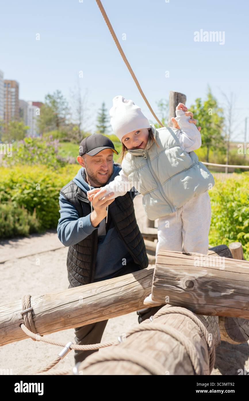 Un padre sostiene sua figlia mentre si bilancia su una struttura di legno in un parco. Si godono una giornata di sole all'aperto, sorridono e spendono qualità Foto Stock