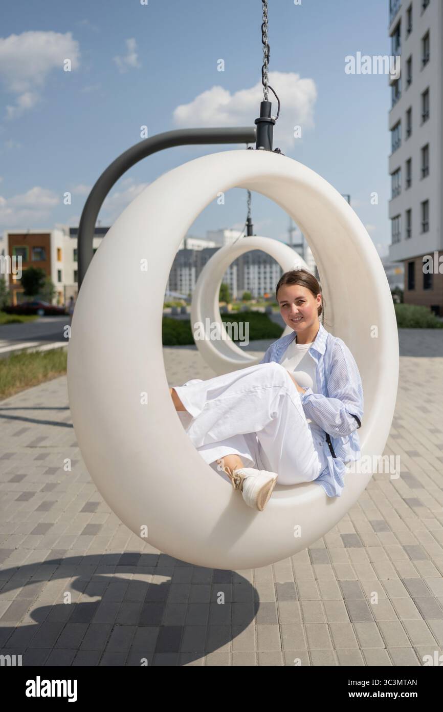 Un'artista donna siede su un moderno altalena circolare in un parco cittadino, godendo di un momento tranquillo di creatività e relax. La giornata di sole mette in risalto la u Foto Stock