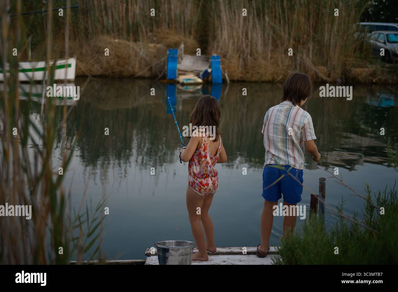 Due bambini si stagliano su un molo sul lago, impegnati a pescare in una giornata tranquilla. Circondati da canne e acque calme, gettano le loro linee nel seren Foto Stock