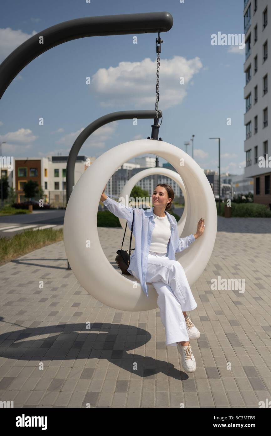 Un'artista donna siede su un moderno altalena circolare in un parco cittadino, godendo di un momento tranquillo di creatività e relax. La giornata di sole mette in risalto la u Foto Stock