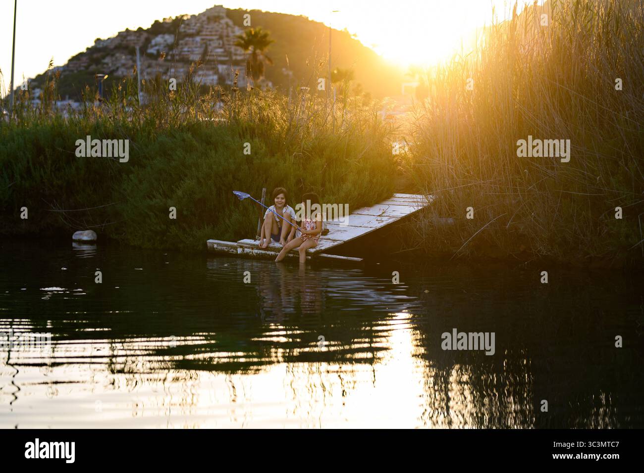 Due bambini siedono su un molo di legno vicino all'acqua, godendosi una tranquilla giornata di pesca al tramonto. Circondati dalla natura, immergono i piedi nell'acqua, cre Foto Stock