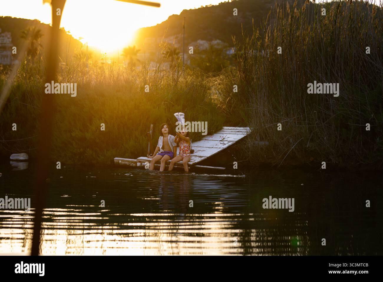 Due bambini potranno divertirsi a pescare in un lago, circondato da alte canne e dal caldo bagliore del sole che tramonta. Tranquillità e natura si fondono in pe Foto Stock