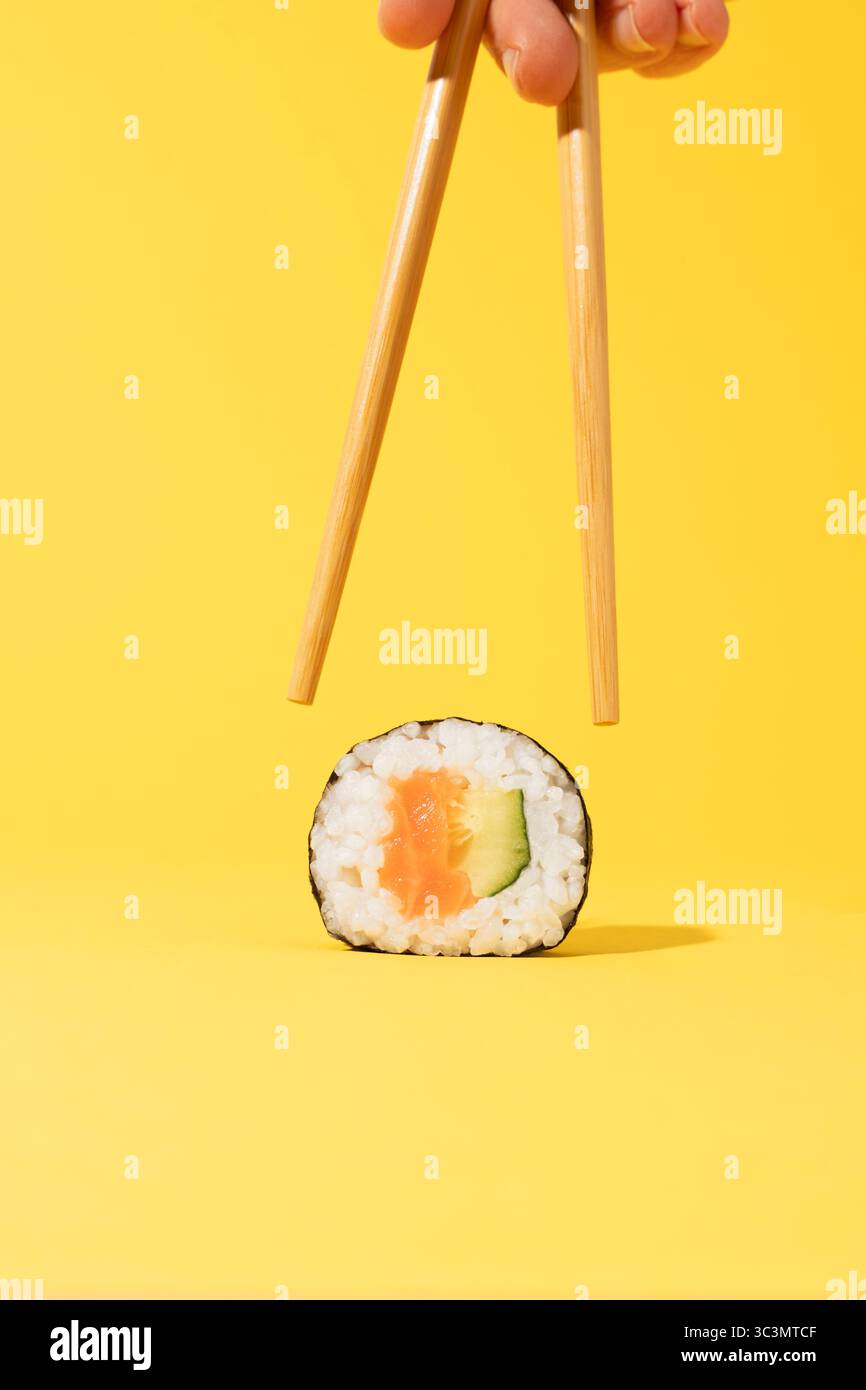 Persona corta e irriconoscibile che tiene le bacchette vicino al sushi roll con salmone, cetriolo e riso su uno sfondo giallo brillante, evidenziando il Foto Stock