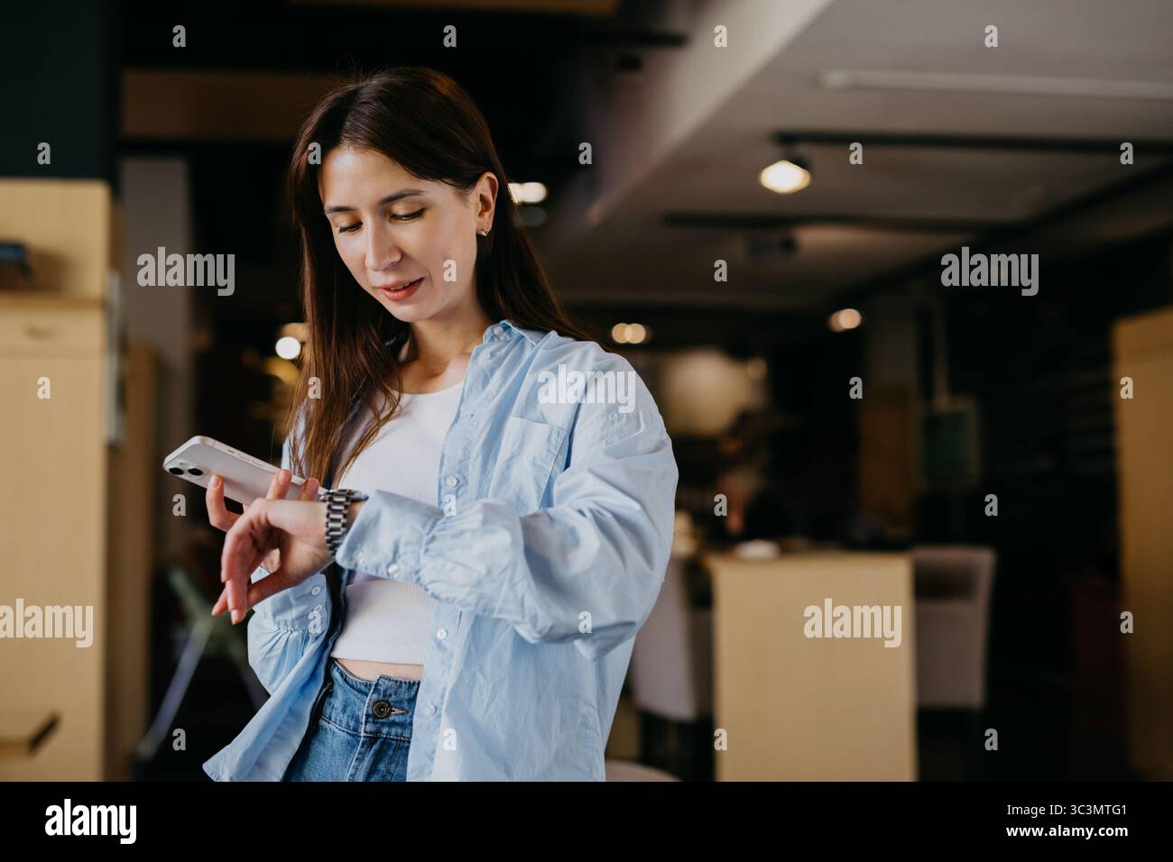 Una donna che indossa una camicia blu controlla il suo orologio mentre tiene in mano uno smartphone in un ambiente accogliente. Sembra concentrata, gestendo il suo tempo e il suo lavoro in modo scheggiato Foto Stock