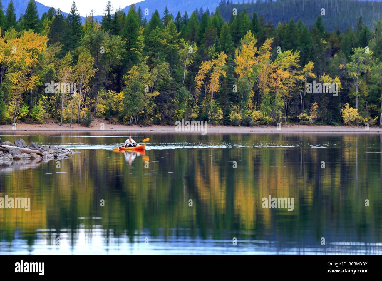 Un kayak solitario scivola attraverso la superficie vetrata del lago McDonald, incorniciata da riflessi della foresta autunnale e da lontane vette montane sotto un cielo ripido. Foto Stock