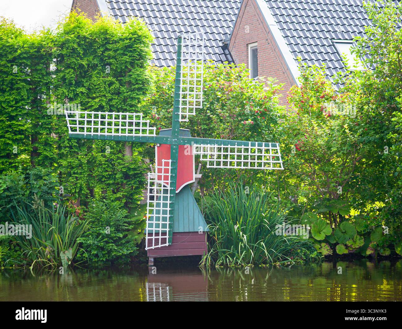 Rustico mulino a vento in scala olandese circondato da vegetazione lussureggiante e un tranquillo corso d'acqua riflette il tranquillo fascino rurale. Foto Stock