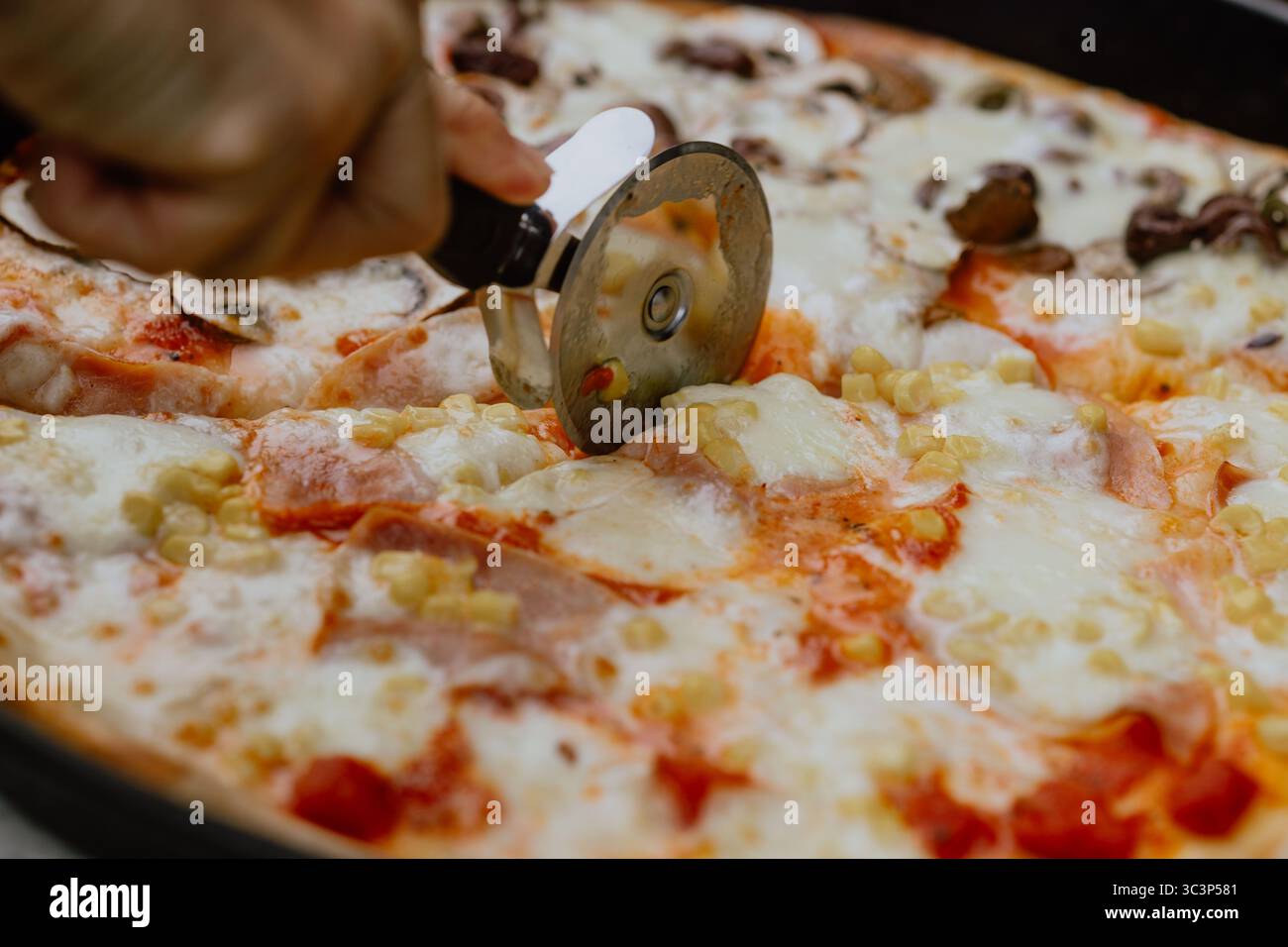 Tagliare la pizza fresca cotta nel forno a legna con mozzarella, funghi e prosciutto utilizzando un rullo per pizza Foto Stock