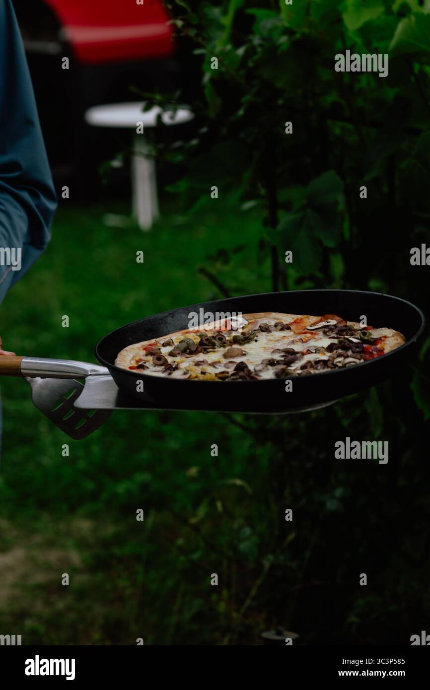 Pizza cotta a legna appena sfornata su un vassoio di metallo con sfondo verde rustico Foto Stock