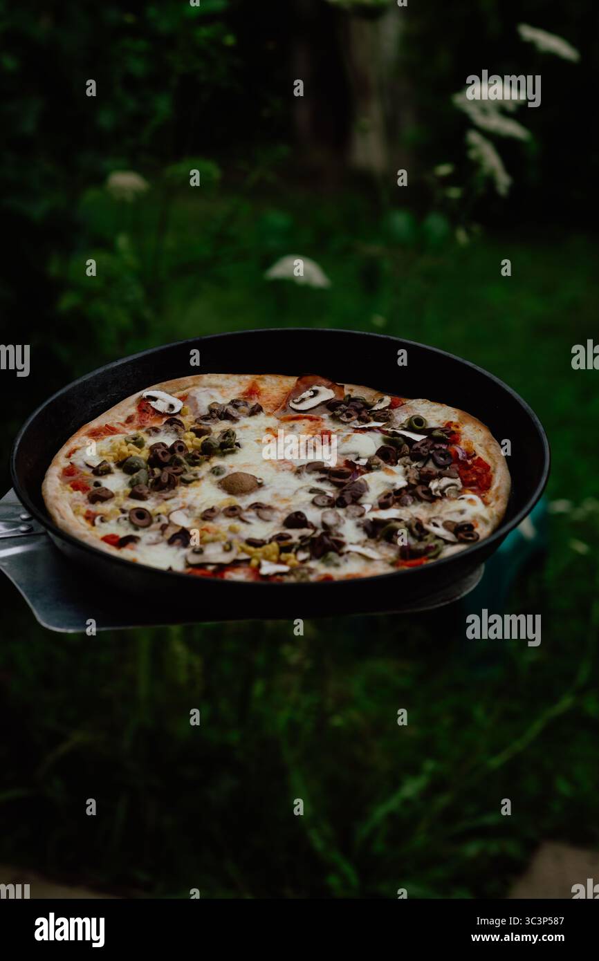Pizza cotta a legna appena sfornata su un vassoio di metallo con sfondo verde rustico Foto Stock