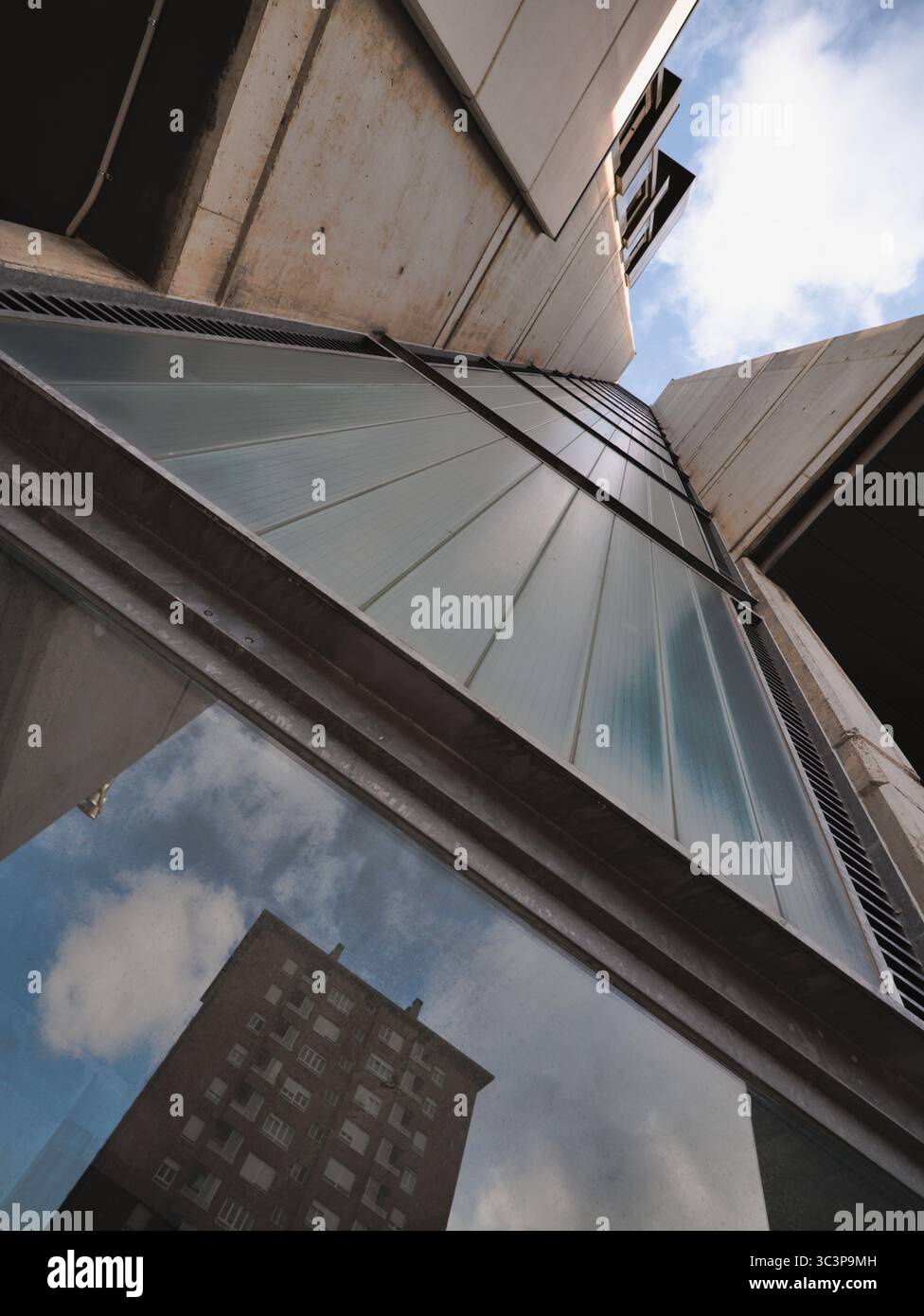 Facciata in cemento di un edificio residenziale con sostegni verticali, inclinati verso l'alto. La struttura è caratterizzata da pannelli in vetro allungati che riflettono il cielo nuvoloso Foto Stock