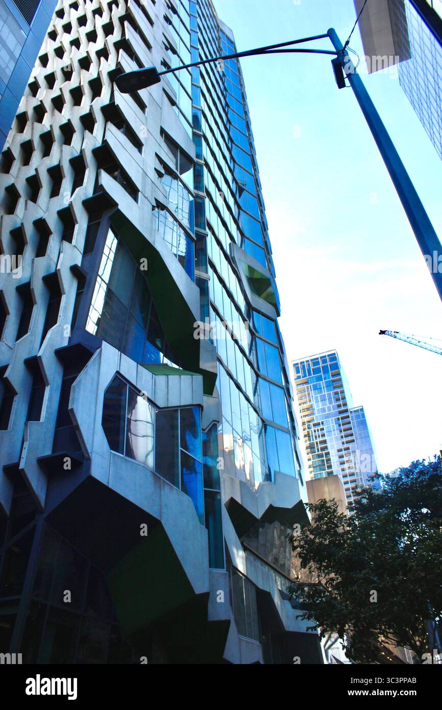 Scena di strada a Melbourne, Australia. Blocco di uffici con finestre di vetro blu che creano interessanti prospettive con alberi, gru e lampioni. Foto Stock