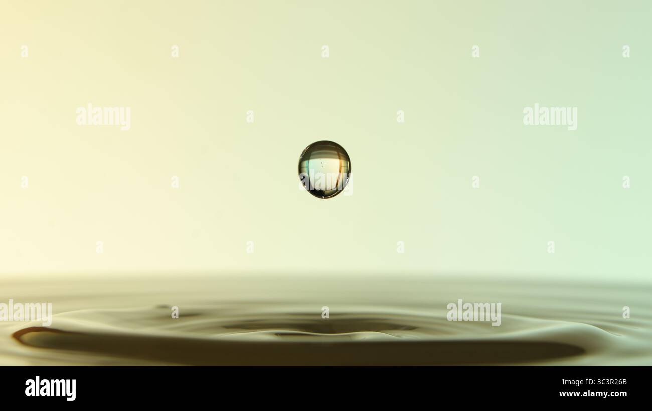 Primo piano di gocce d'acqua sospese su una superficie calma, che formano increspature e riflessi su sfondi a gradiente morbido. Foto Stock