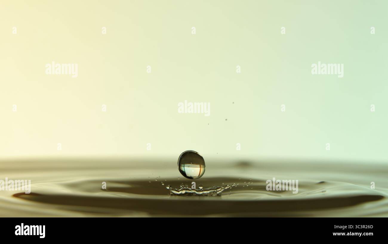 Primo piano di gocce d'acqua sospese su una superficie calma, che formano increspature e riflessi su sfondi a gradiente morbido. Foto Stock