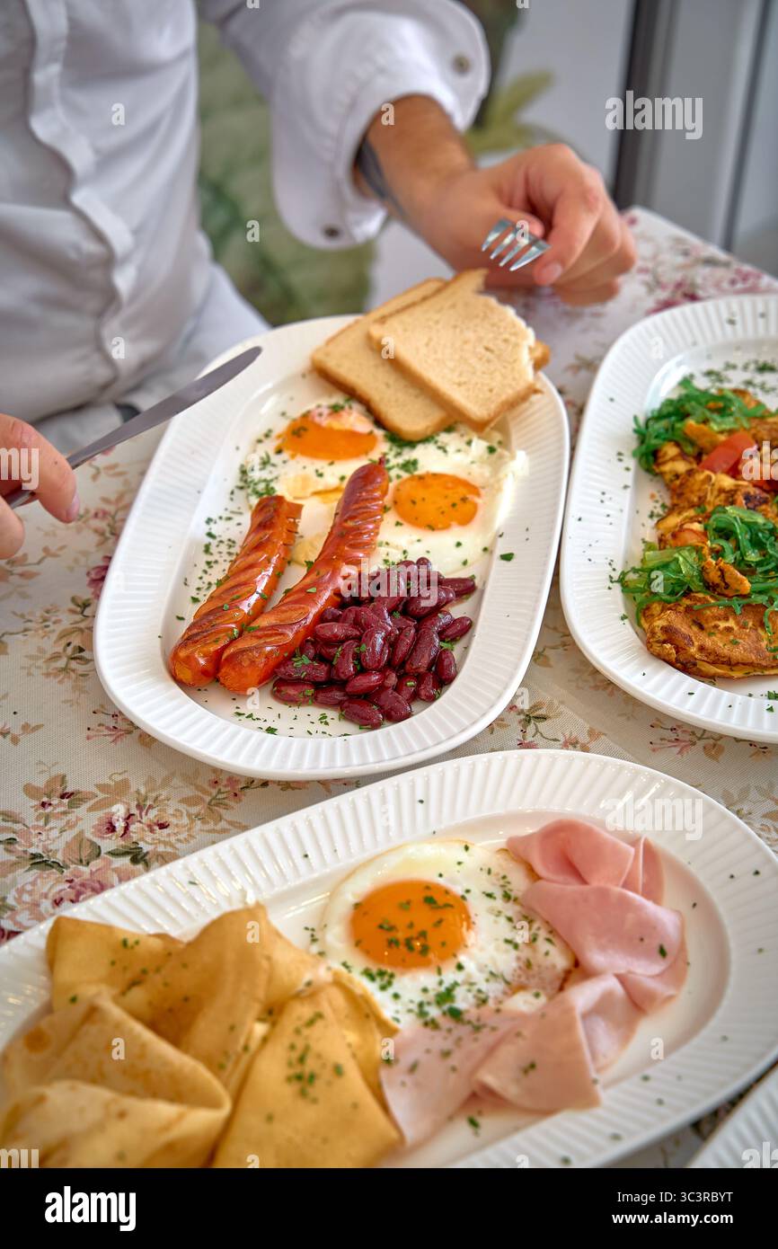 Una deliziosa colazione a buffet con uova, salsicce, fagioli renali, prosciutto e crepes su una tovaglia floreale per un delizioso pasto mattutino Foto Stock