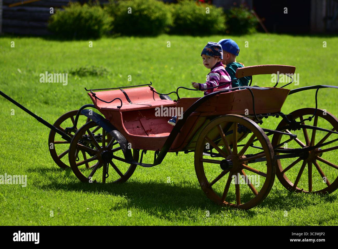 Due bambini si godono un giro in un vagone rosso vintage in legno su un prato erboso soleggiato. Fascino rurale, amicizia e ricordi di infanzia felici in un ambiente tranquillo Foto Stock