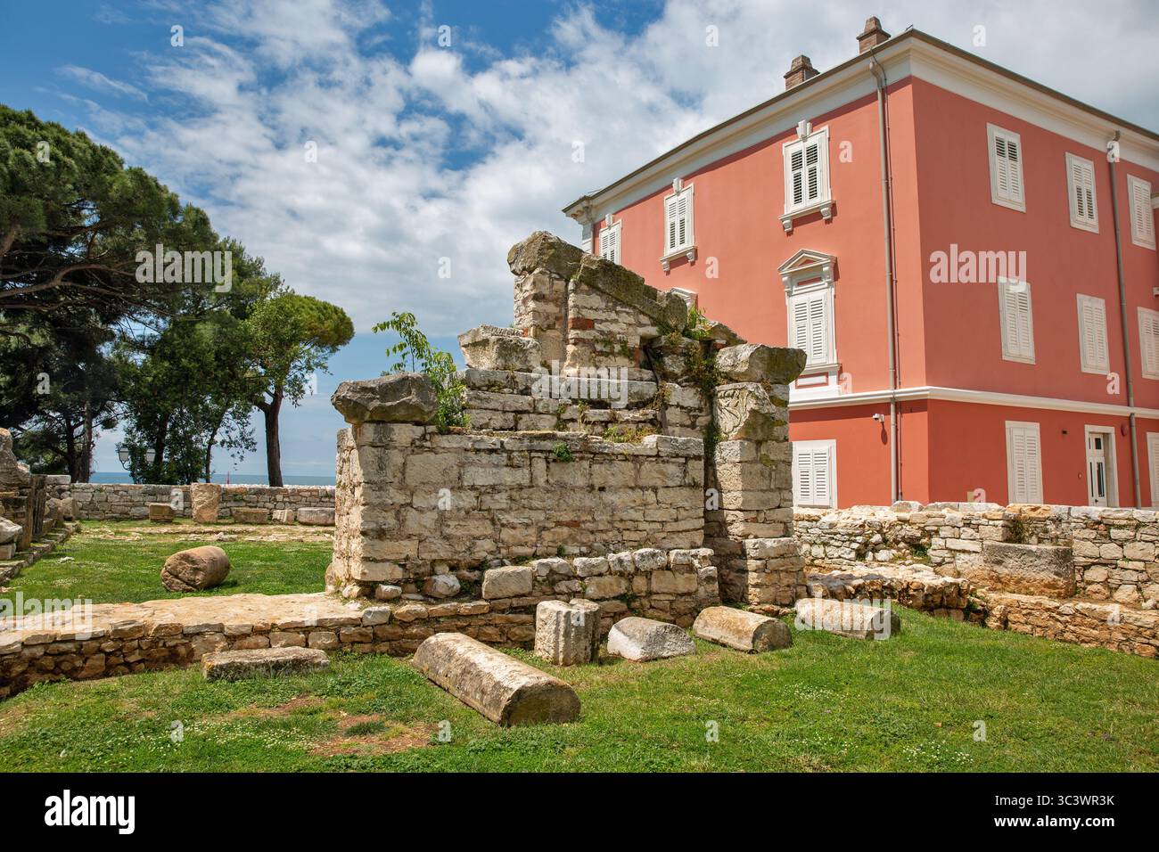 Antiche rovine romane in pietra e resti archeologici accanto all'elegante edificio rosa con persiane bianche a Porec, Croazia, che mostra il contrasto tra una Foto Stock