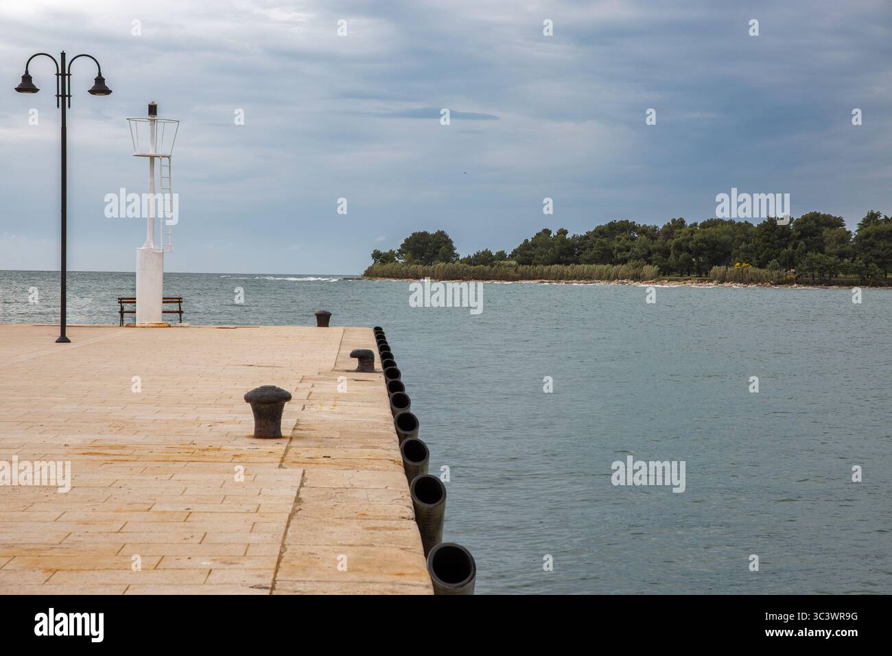 Tranquillo molo in pietra con faro bianco, lampioni e dissuasori che si affacciano sulle calme acque dell'Adriatico e sulla costa boscosa di Novigra Foto Stock
