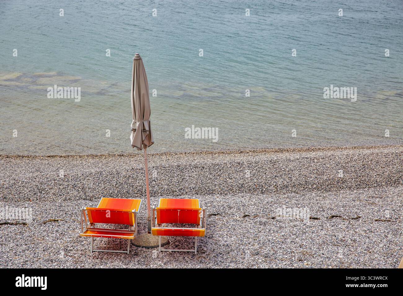Due sedie arancioni luminose e ombrellone chiuso riposano su una tranquilla spiaggia di ciottoli in Croazia, un luogo sereno e romantico per due vicino alle dolci onde dell'Adriatico Foto Stock
