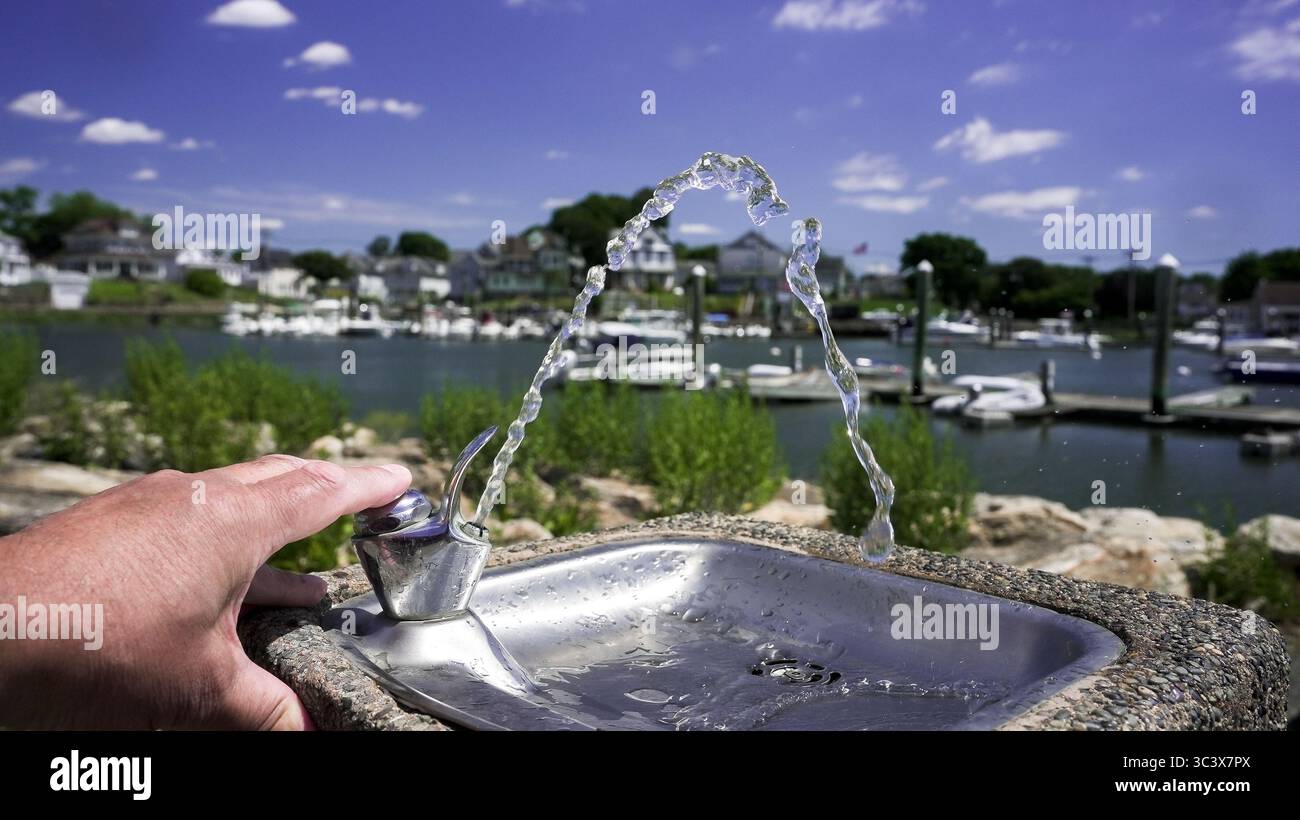 Una persona che si accende spingendo una fontana d'acqua con uno sfondo di barche attraccate in un tranquillo canale navigabile e un cielo azzurro limpido, creando un bancomat rinfrescante Foto Stock