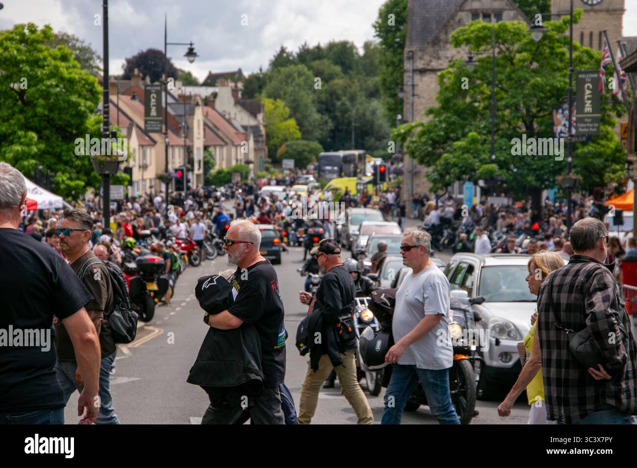 Una vivace scena di strada con persone che partecipano a un evento motociclistico nella città di Calne Wiltshire nel Regno Unito, circondata da veicoli parcheggiati e lussureggianti alberi verdi Foto Stock