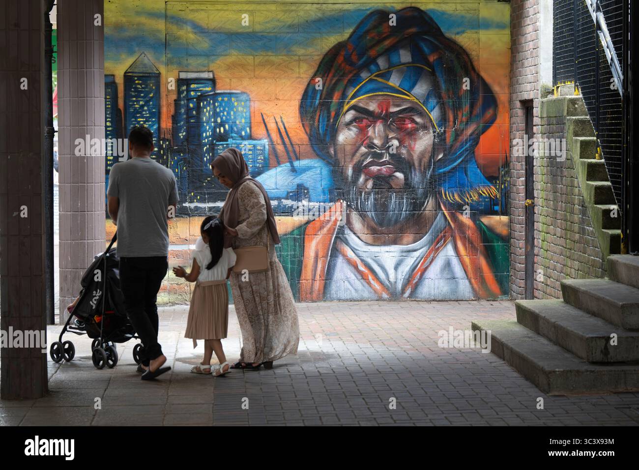 Una famiglia passeggia davanti a un murale raffigurante una figura turbanata e lo skyline di Canary Wharf a Poplar, Londra. Il murale aggiunge colore al paesaggio urbano. Foto Stock